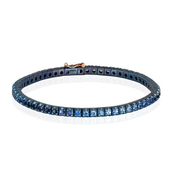Muška klasična titanijumska tenis narukvica od anodizovanog plavog Titanijuma sa safirima u nijansama plave boje. Narukvica se sastoji od neprekidnog niza Safira, svaki težine 0,05ct, postavljenih na titanijumske segmente, smeštenih jedan pored drugog. Kopča je sastavljena od dugmeta sa jezičkom na kojem je postavljen safir i sigurnosnog osigurača od 18 kt roze zlata. Ova narukvica kombinuje ideju klasičnog tenis nakita sa inovacijom materijala, čineći je laganom, sigurnom i lakom za nošenje.
