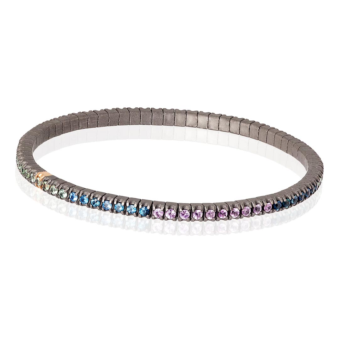 Muška narukvica od titanijuma, sa rainbow safirima model tenbrtzamulrg3pt_p-2 Muška elastična tenis narukvica od titanijuma i višebojnih safira (2,69 ct). Narukvica se sastoji od neprekidnog niza kamenja težine po 0,03 tačke postavljenih na titanijumske segmente, sa jednim kamenom ufasovanim u 18 kt roze zlato. Svaki se nalazi jedan pored drugog na elastičnoj vezi. Ova narukvica spaja ideju klasičnog tenis nakita sa inovacijom materijala, čineći je laganom, sigurnom i lakom za nošenje.
