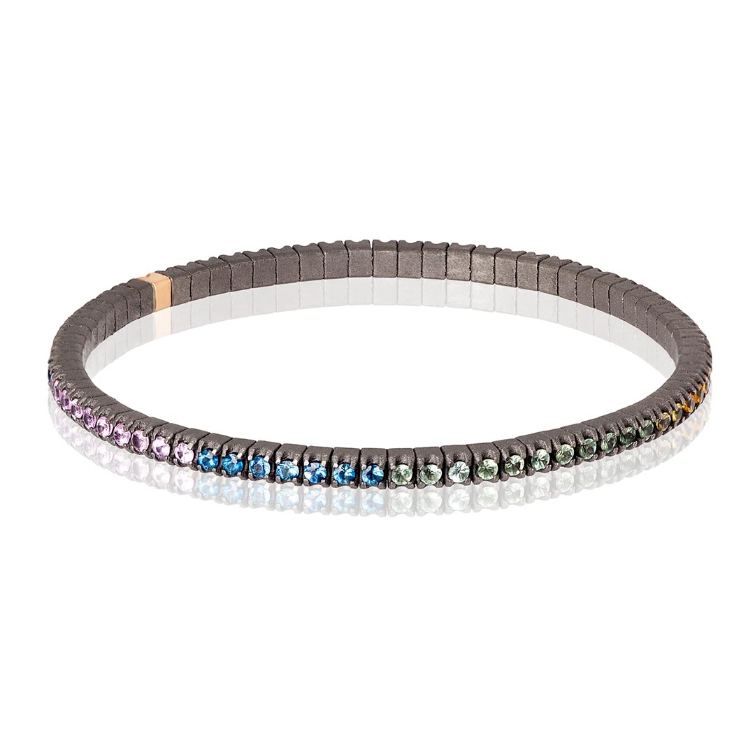 Muška narukvica od titanijuma, sa rainbow safirima model tenbrtzamulrg3pt_p-3 Muška elastična tenis narukvica od titanijuma i višebojnih safira (2,69 ct). Narukvica se sastoji od neprekidnog niza kamenja težine po 0,03 tačke postavljenih na titanijumske segmente, sa jednim kamenom ufasovanim u 18 kt roze zlato. Svaki se nalazi jedan pored drugog na elastičnoj vezi. Ova narukvica spaja ideju klasičnog tenis nakita sa inovacijom materijala, čineći je laganom, sigurnom i lakom za nošenje.