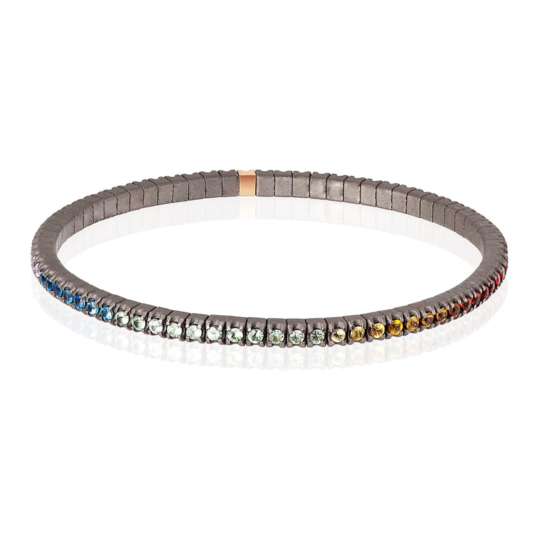 Muška narukvica od titanijuma, sa rainbow safirima model tenbrtzamulrg3pt_p-4 Muška elastična tenis narukvica od titanijuma i višebojnih safira (2,69 ct). Narukvica se sastoji od neprekidnog niza kamenja težine po 0,03 tačke postavljenih na titanijumske segmente, sa jednim kamenom ufasovanim u 18 kt roze zlato. Svaki se nalazi jedan pored drugog na elastičnoj vezi. Ova narukvica spaja ideju klasičnog tenis nakita sa inovacijom materijala, čineći je laganom, sigurnom i lakom za nošenje.