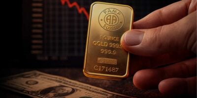 Ruka drži pamp zlatnu polugu investicionog zlata iznad dolara na stolu i grafikona koji pokazuje crvenu liniju nadole